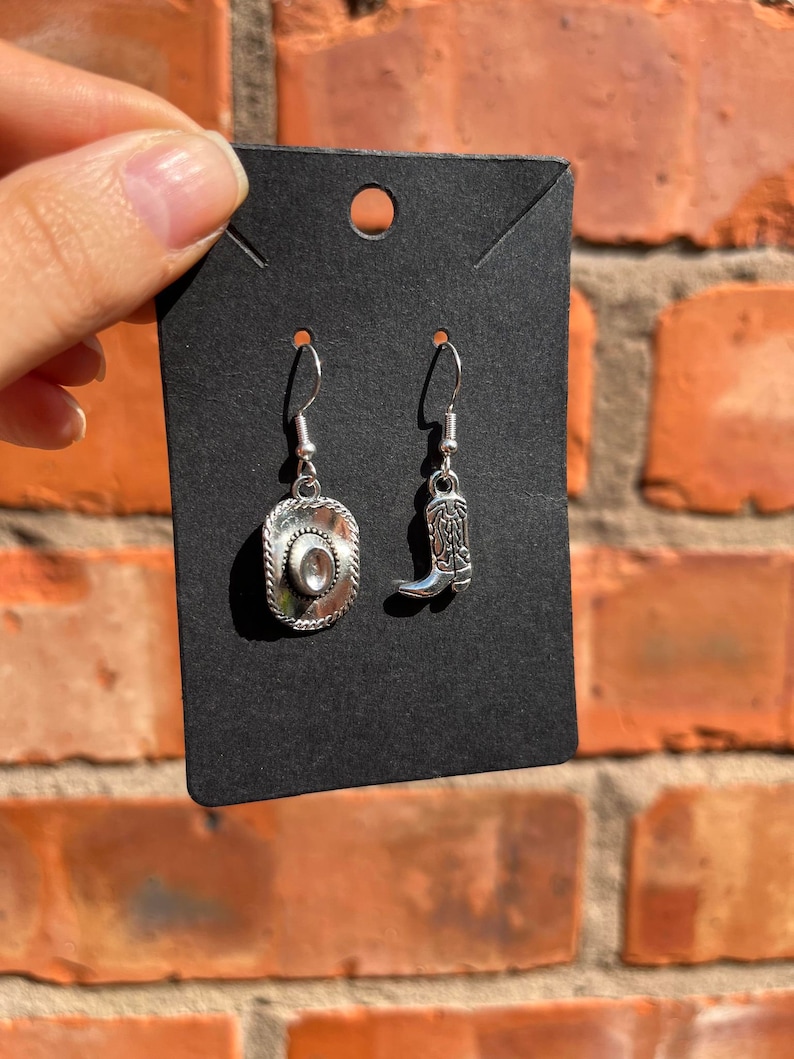 May include: Silver-toned cowboy-themed earrings. One earring is a cowboy hat, the other a cowboy boot. The earrings are displayed on a black card against a brick background.