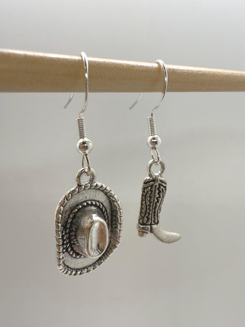 May include: Silver-toned cowboy hat and boot dangle earrings. The hat has a detailed brim and crown, while the boot features intricate embossed patterns. The earrings hang from silver-colored fish hook ear wires, perfect for a Western-themed look.