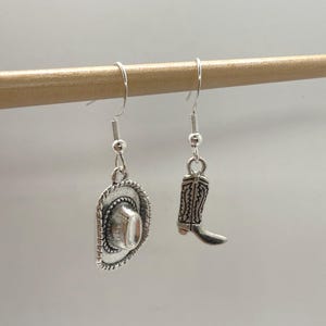 May include: Silver-toned cowboy hat and boot dangle earrings. The hat has a detailed brim and crown, while the boot features intricate stitching. These earrings are a charming accessory for a Western-themed look.