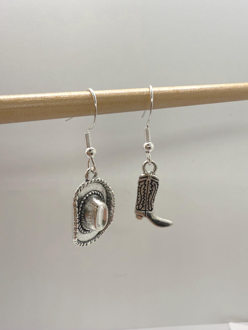 May include: Silver-toned cowboy hat and boot dangle earrings. The hat has a detailed brim and crown, while the boot features intricate stitching. These earrings are a charming accessory for a Western-themed look.