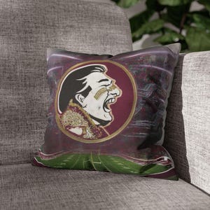 May include: A maroon and gold throw pillow with a vintage-style illustration of a person's face with a Seminole Tribe logo. The background is a blurred image of a football field.