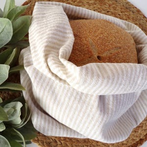 Peut inclure: Un pain rond dans un sac à pain en lin rayé crème et beige. Le pain repose sur un tapis circulaire tissé. Des brins de feuilles vertes sont visibles à gauche. Le sac à pain est en lin.