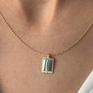 May include: A gold chain necklace with a small, rectangular pendant. The pendant is smooth and has a slightly reflective surface.