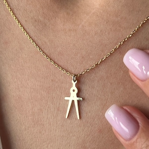 May include: A gold chain necklace with a compass pendant. The necklace is worn by a person with a white shirt and pink nail polish.