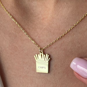 May include: A gold chain necklace with a pendant shaped like a box of french fries. The pendant has the word "CHIPS" inscribed on it.