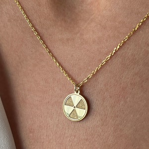 Puede incluir: Un collar de cadena dorada con un colgante circular que presenta un símbolo de radiación blanco.