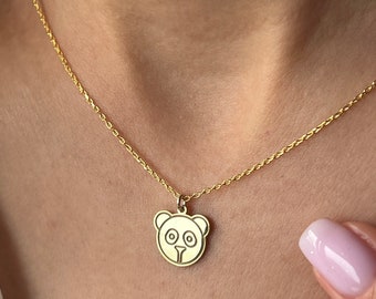 May include: A gold-colored necklace featuring a pendant shaped like a bear's face. The bear pendant has circular eyes and a small nose. The necklace is worn against a white shirt.