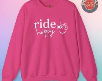 Op de afbeelding: Roze sweatshirt met witte tekst die "ride happy" zegt en een witte fietsafbeelding met een smiley.