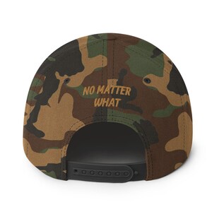 May include: Camouflage baseball cap with the embroidered text "NO MATTER WHAT" in a gold color. The cap features a black adjustable snapback closure. The hat's pattern includes shades of brown, green, and black.