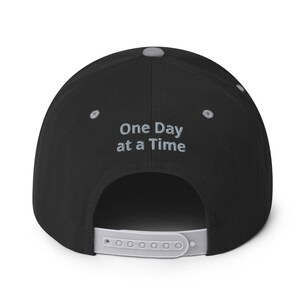 May include: Black baseball cap with a gray brim and the text "One Day at a Time" embroidered in gray on the back.