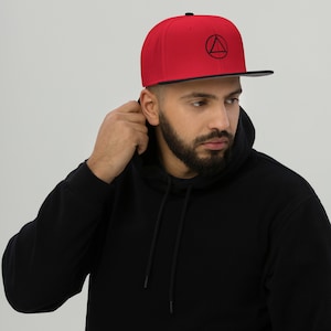 May include: A red and black baseball cap with a black circle and triangle design. The cap has a flat brim and a black strap.