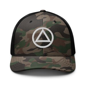 May include: Camouflage trucker hat with a white triangle within a circle emblem on the front. The hat features a black mesh back and a curved brim. The camouflage pattern includes shades of green, brown, and tan.