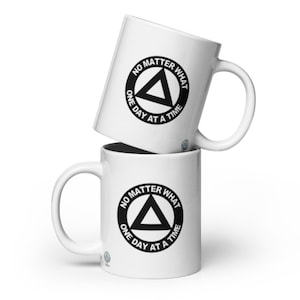 May include: Two white ceramic mugs stacked on top of each other. Each mug features a black circular design with a triangle and the text "NO MATTER WHAT ONE DAY AT A TIME". The mugs have a curved handle.