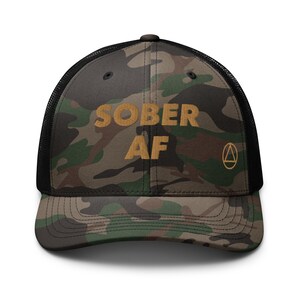 May include: A black and green camouflage trucker hat with the text "SOBER AF" in gold embroidery. The hat also features a small gold triangle symbol.