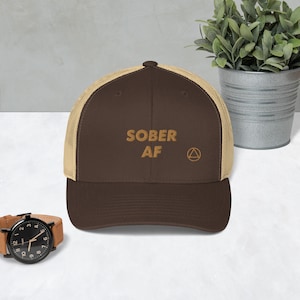 May include: A brown and beige trucker hat with the text "SOBER AF" and a yellow triangle symbol. The hat has a mesh back.