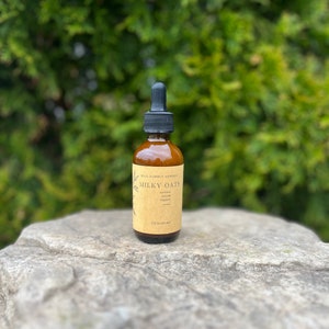 May include: A brown glass bottle with a black dropper, labeled "Milky Oats" with text below. The bottle contains a liquid and is 2 fl oz (60 ml). The bottle is on a gray rock with a green leafy background.