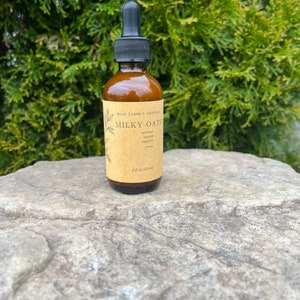 May include: A brown glass bottle with a black dropper, labeled "Milky Oats" with text below. The bottle contains a liquid and is 2 fl oz (60 ml). The background is a green bush, and the bottle sits on a gray rock.