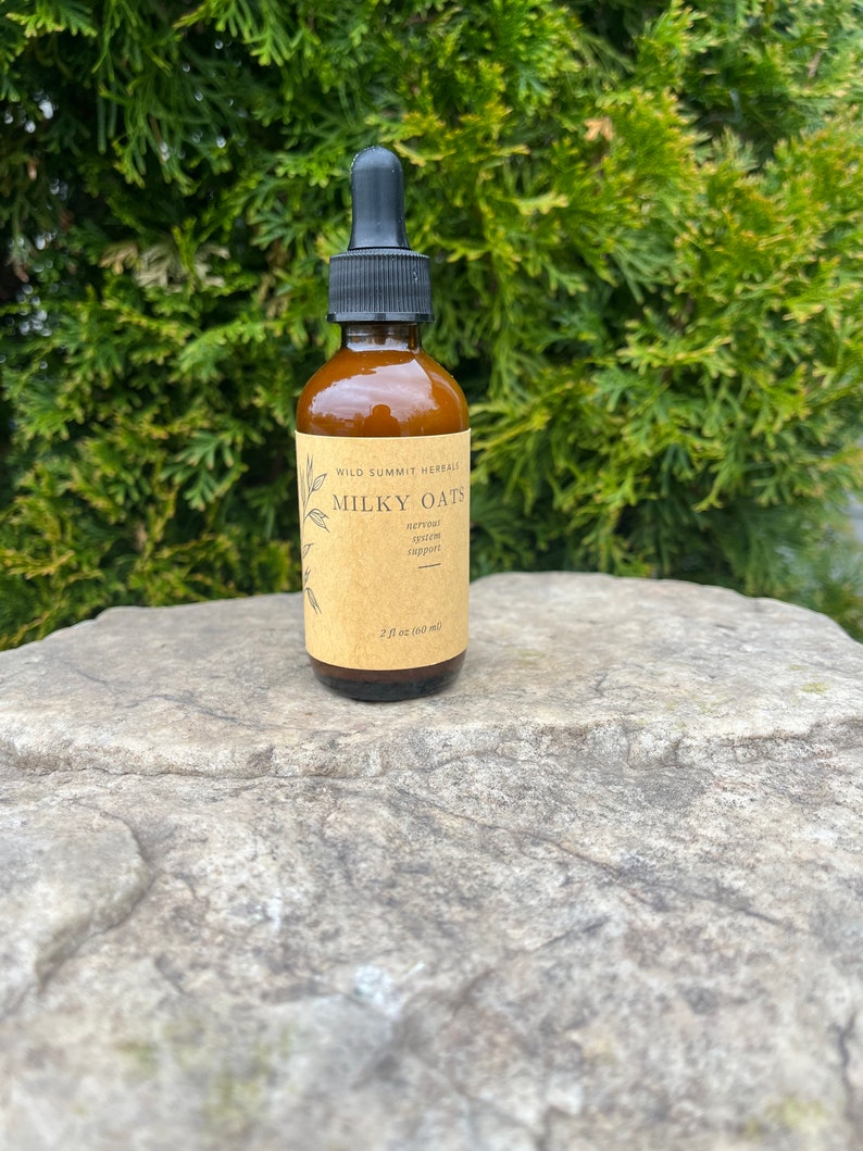May include: A brown glass bottle with a black dropper, labeled "Milky Oats" with text below. The bottle contains a liquid and is 2 fl oz (60 ml). The background is a green bush, and the bottle sits on a gray rock.