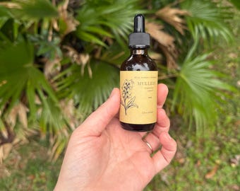 May include: A brown glass bottle with a black dropper, labeled "MULLEIN" with botanical illustrations. The label is a light brown color. The bottle is held in a hand against a backdrop of green foliage.