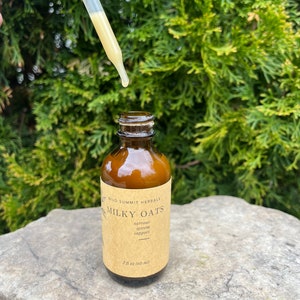 May include: A brown glass bottle with a label reading "Milky Oats" and "nervous system support." A dropper dispenses a yellow liquid. The bottle is 2 fl oz (60 ml). The background is a green bush.