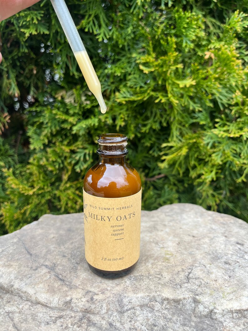 May include: A brown glass bottle with a label reading "Milky Oats" and "nervous system support." A dropper dispenses a yellow liquid. The bottle is 2 fl oz (60 ml). The background is a green bush.