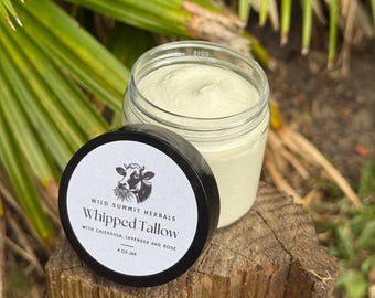 May include: A clear glass jar filled with pale green whipped tallow, featuring a black lid with a white label. The label displays a cow illustration, the text Wild Summit Herbals, Whipped Tallow, with calendula, lavender and rose, and 4 oz jar.