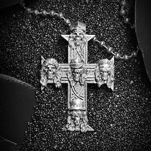 Puede incluir: Un colgante de cruz de plata con cuatro caras de calavera, cada una con un peinado diferente, sobre un fondo negro.