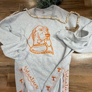 May include: Light gray sweatshirt with an orange hound dog graphic. The sleeves have the word "Volunteers" and the letter "T" embroidered in orange. A wooden bead garland and greenery are in the background.