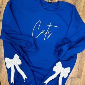 May include: A royal blue sweatshirt with white embroidered bows on the sleeves and the word "Cats" in white script on the front.