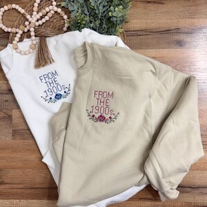 Op de afbeelding: Twee crewneck sweatshirts, één crème en één beige, worden getoond. Elk heeft de geborduurde tekst "FROM THE 1900s" met bloemmotieven. De sweatshirts liggen op een houten oppervlak, met decoratieve kralen en groen in de achtergrond.