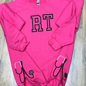 May include: Pink sweatshirt with black embroidered letters "RT" and two black and white stethoscope designs on the sleeves.