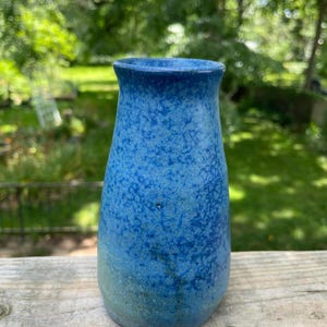 May include: A handmade ceramic vase with a speckled blue glaze. The vase has a rounded body and a slightly flared rim. The colour transitions from a lighter blue-green at the base to a deep blue at the top. The vase is displayed on a wooden surface.