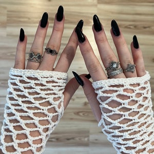 May include: A pair of white crocheted fingerless gloves with black nail polish on the fingers. The gloves have a delicate, lacy pattern and are adorned with silver rings.