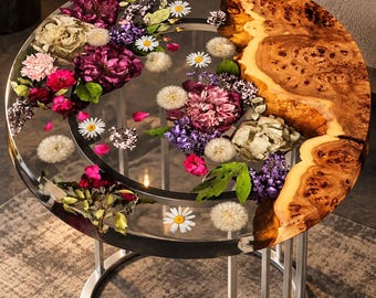 May include: A round side table featuring a clear resin tabletop embedded with various dried flowers, including purple peonies, white daisies, and dandelions, alongside a natural wood burl edge and a silver-colored metal frame base.