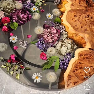 May include: A round table top made of wood and resin with a clear epoxy resin layer. The resin layer contains pressed flowers and leaves in shades of pink, purple, white, and green. The wood portion of the table top is a natural brown color with a unique grain pattern.