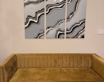 Puede incluir: Un conjunto de arte mural abstracto de tres piezas con un diseño fluido en plata, blanco, negro y dorado. Debajo, un sofá de terciopelo dorado con mechones de canal verticales y patas doradas.