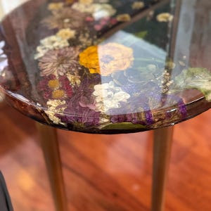 Puede incluir: Una mesa auxiliar redonda con una tapa de resina transparente con flores secas en tonos amarillos, blancos y morados. La mesa tiene tres patas de madera con detalles dorados en la base. El diseño de la mesa es una mezcla única de elementos naturales y estilo moderno.