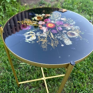 Puede incluir: Una mesa auxiliar redonda con una tapa de cristal negro que presenta un diseño floral en tonos rosa, morado, amarillo y blanco. La mesa tiene una estructura y patas de metal dorado, creando una pieza decorativa elegante. Diámetro aproximado de 45 cm.