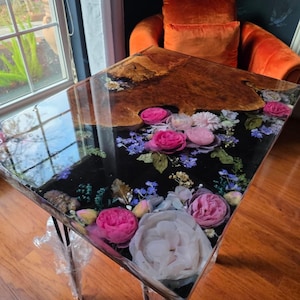 Puede incluir: Mesa cuadrada de resina con tapa de madera y arreglo floral. La mesa presenta una superficie de resina transparente con flores rosas y blancas incrustadas, detalles azules y un fondo oscuro. Las patas son transparentes.