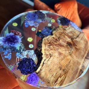 Puede incluir: Mesa auxiliar redonda con tapa de resina transparente con flores azules y moradas incrustadas. La mesa presenta un borde de madera natural y descansa sobre patas plateadas. Una pieza única para la decoración del hogar, aprox. 60 cm de diámetro.