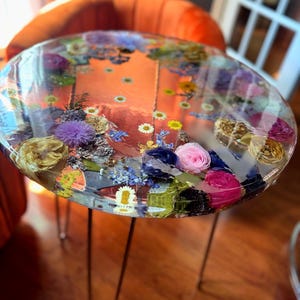Puede incluir: Mesa redonda de resina transparente con flores secas incrustadas en tonos rosa, amarillo, azul y morado. La mesa tiene tres patas finas de metal. El fondo incluye una silla naranja y un suelo de madera.