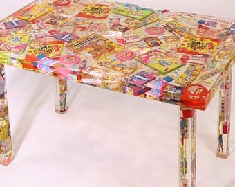 May include: A clear acrylic table with a colorful candy-themed design. The table top is covered in a variety of candy wrappers and logos, including brands like Skittles, Starburst, and Nerds. The table has four clear acrylic legs.