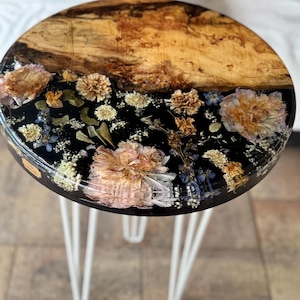 Puede incluir: Una mesa auxiliar redonda con tapa de madera y patas de horquilla blancas. La parte superior es mitad madera natural y mitad resina negra con flores secas incrustadas en tonos beige, rosa y azul. La mesa tiene unos 46 cm de diámetro.