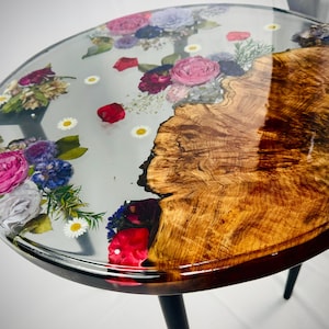 May include: Round coffee table with a clear resin top inlaid with colorful flowers and a natural wood grain base. The table has a dark rim and black legs. The flowers include pink roses, red petals, and white daisies.