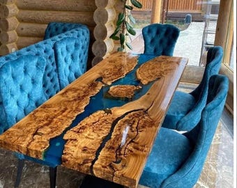 May include: A rectangular wooden dining table with a blue epoxy river design. The table is surrounded by blue upholstered chairs. The wood has a natural grain pattern, and the epoxy creates a contrasting blue river effect.