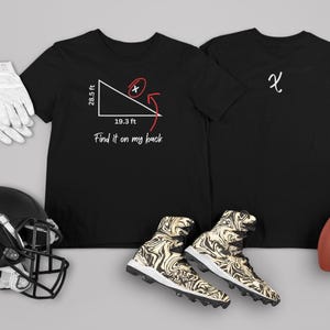 Peut inclure: Deux t-shirts noirs, l'un avec un graphique blanc d'un triangle rectangle avec le texte "Find it on my back" et l'autre avec une lettre blanche "X". Les t-shirts sont disposés sur une surface grise avec un ballon de football, un casque de football et une paire de gants de football blancs.
