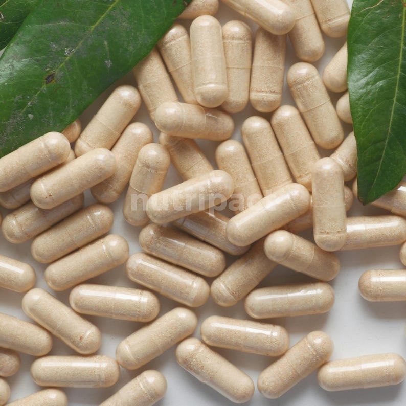 Puede incluir: Primer plano de numerosas c&aacute;psulas beige, posiblemente suplementos, dispuestas sobre una superficie. Las c&aacute;psulas son cil&iacute;ndricas con extremos redondeados. Se ven hojas verdes, aportando un elemento natural. La imagen est&aacute; bien iluminada.