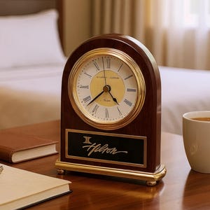 May include: A wooden clock with a gold-colored face and Roman numerals. The clock has a dark brown wooden frame with gold accents and the Hilton logo. A cup of coffee, a notebook, and a pen are on the table.