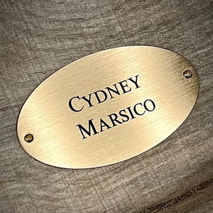 May include: An oval-shaped brass nameplate with a brushed metallic finish. The plate features the engraved black text CYDNEY MARSICO in the center and includes two small circular screws, one on the far left and one on the far right side.