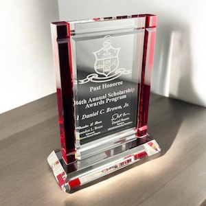 May include: A clear glass award with red accents. The award features the text "Past Honoree" and "14th Annual Scholarship Awards Program". The name "Daniel C. Brown, Jr." is also visible. The award is rectangular with a beveled base.